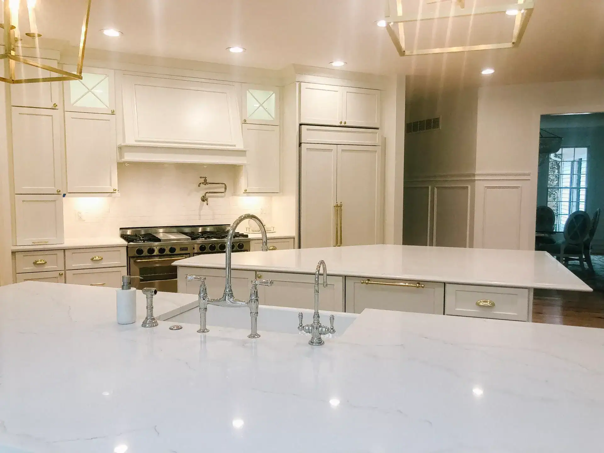 Cream shaker kitchen cabinets with built-in hood, brass hardware, marble-look quartz island and polished chrome bridge faucet
